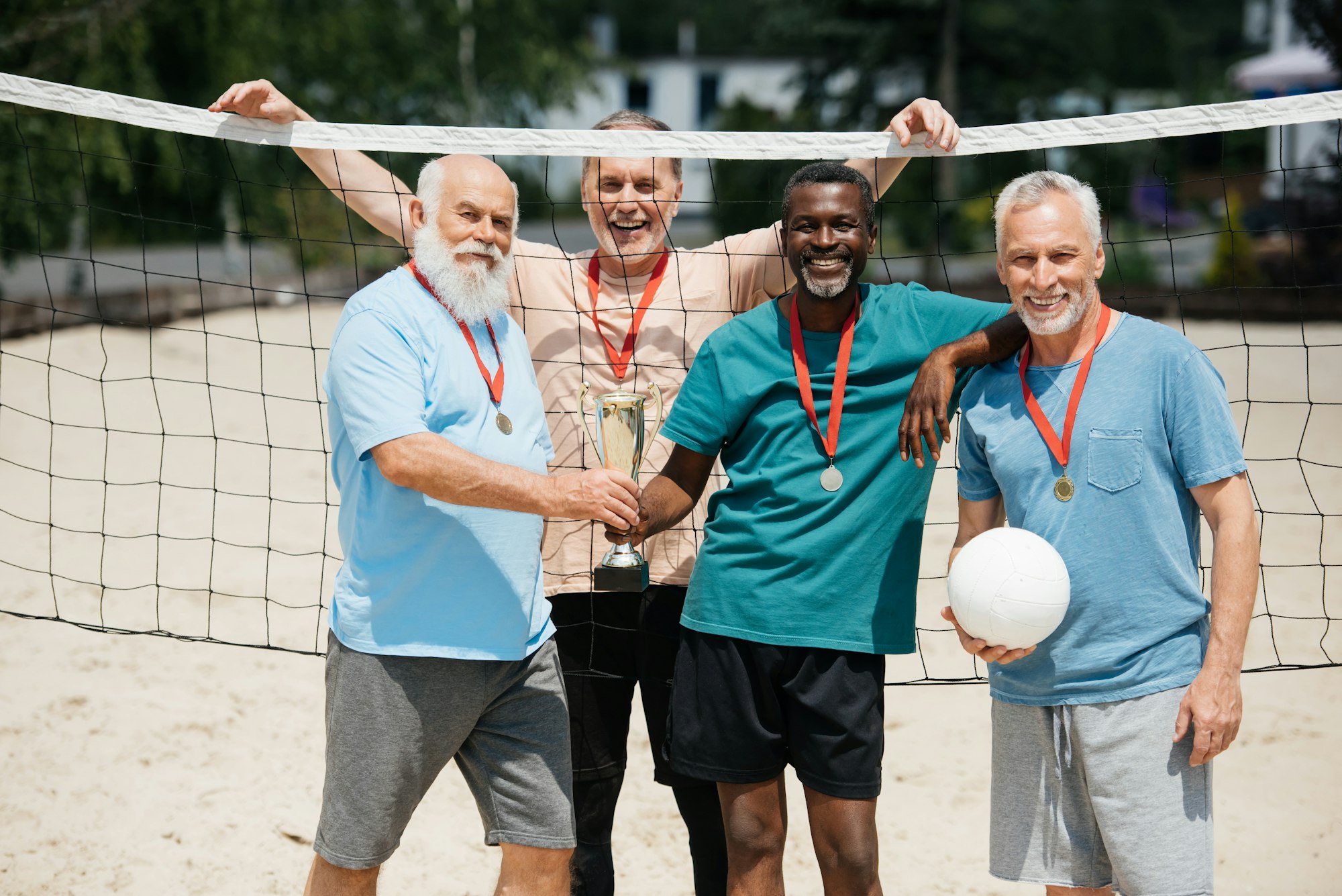 portrait of smiling multiethnic elderly friends with tennis ball, medals and champions cup on beach